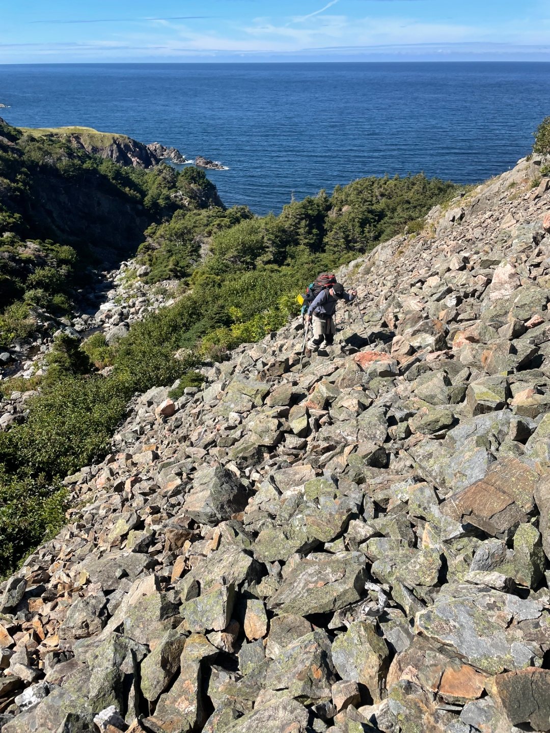 Hiker descending a steep boulder scree slope toward a rocky cove on the Cape Breton coast
